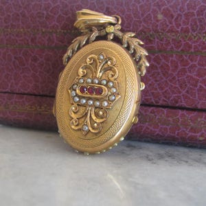 May include: A gold locket with a detailed floral design and a red stone accent. The locket is surrounded by a border of pearls.