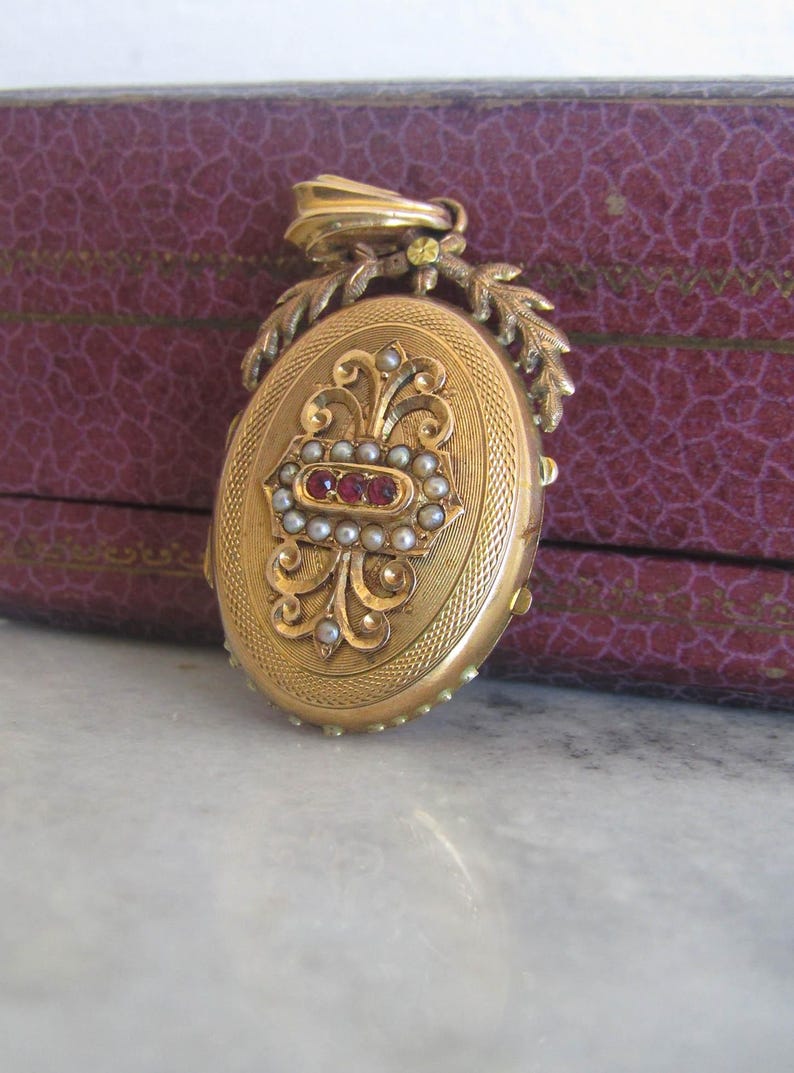 May include: A gold locket with a detailed floral design and a red stone accent. The locket is surrounded by a border of pearls.