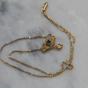 May include: A vintage gold-toned necklace with a decorative pendant featuring a square green gemstone. The pendant has an ornate design, and a small heart charm dangles below. The necklace is displayed on a marble surface.