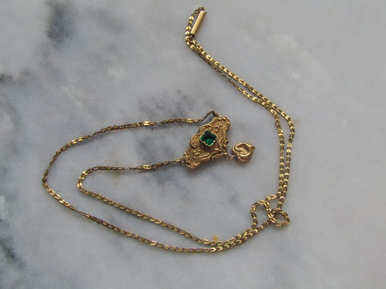 May include: A vintage gold-toned necklace with a decorative pendant featuring a square green gemstone. The pendant has an ornate design, and a small heart charm dangles below. The necklace is displayed on a marble surface.