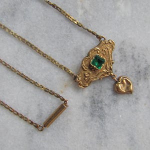 May include: A vintage gold-toned necklace with an ornate pendant featuring a square green gemstone and a heart-shaped charm. The chain is delicate, and the necklace is set against a marble background.