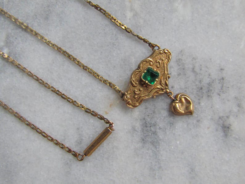 May include: A vintage gold-toned necklace with an ornate pendant featuring a square green gemstone and a heart-shaped charm. The chain is delicate, and the necklace is set against a marble background.