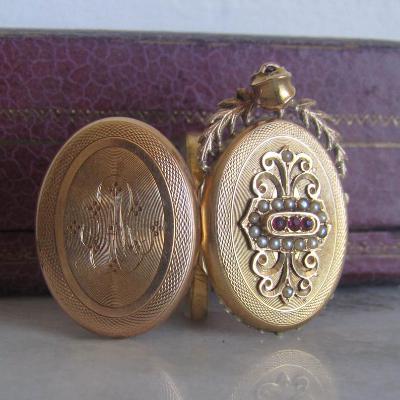 May include: A gold locket with a delicate floral design and a row of red and white gemstones. The locket is open to reveal a gold interior with an engraved monogram.
