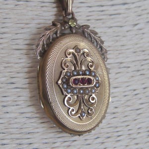 May include: Gold oval locket with a floral design and red and white stones. The locket is on a gold chain.