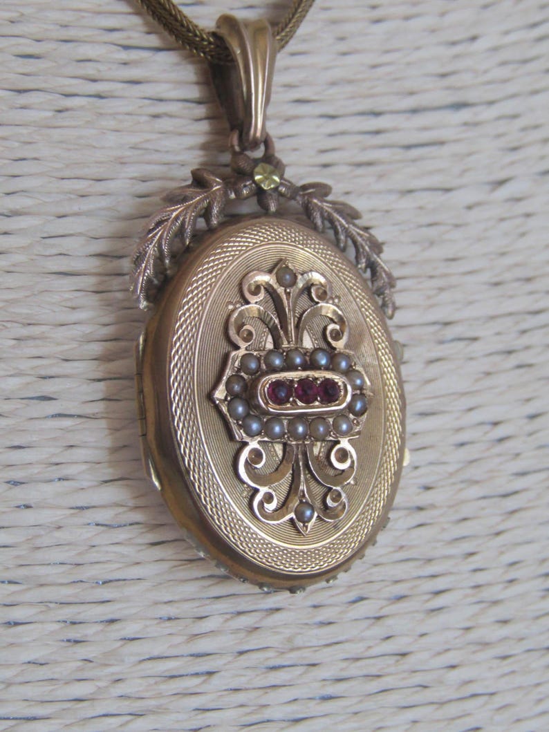 May include: Gold oval locket with a floral design and red and white stones. The locket is on a gold chain.