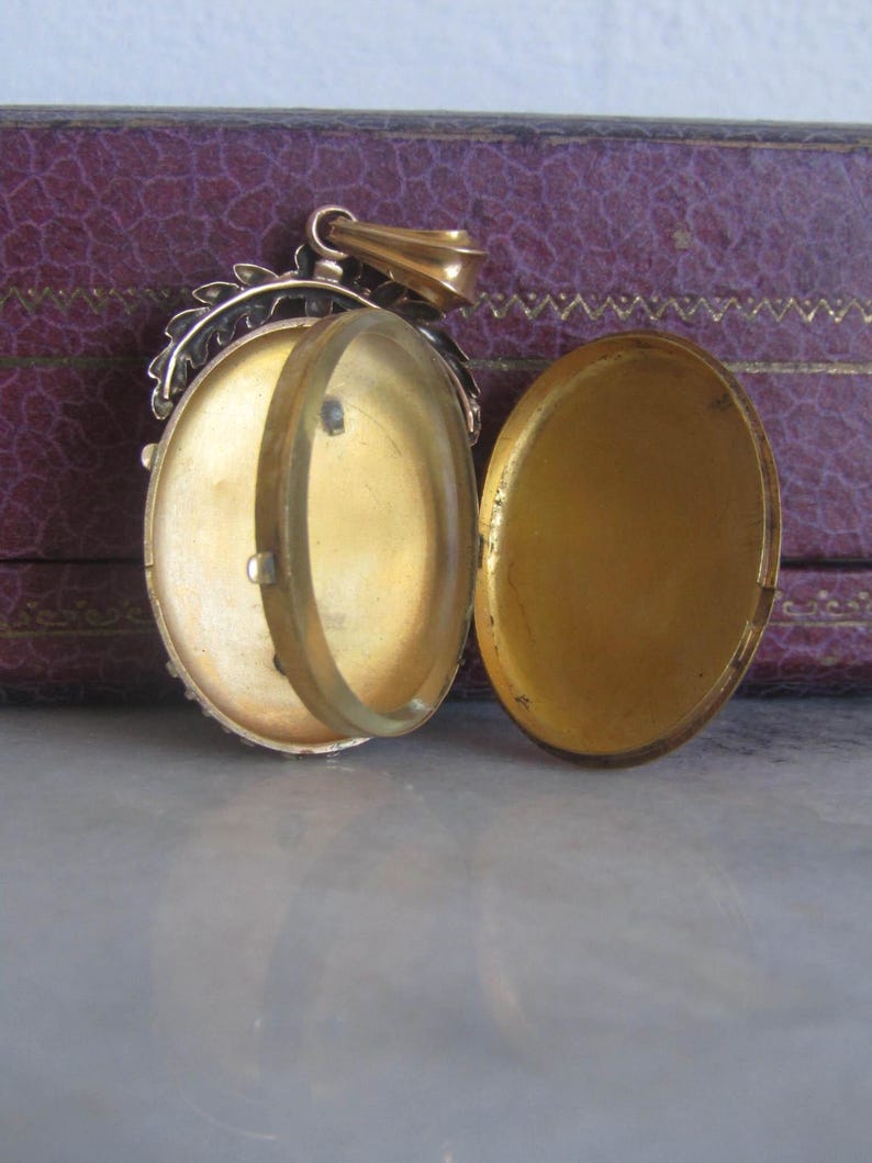 May include: A gold locket with a hinged lid and a decorative floral design around the edge. The locket is open, revealing a clear glass insert.