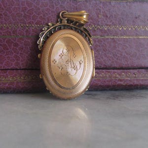 May include: A gold locket with an oval-shaped front and a detailed floral design around the edge. The front of the locket has an engraved monogram.