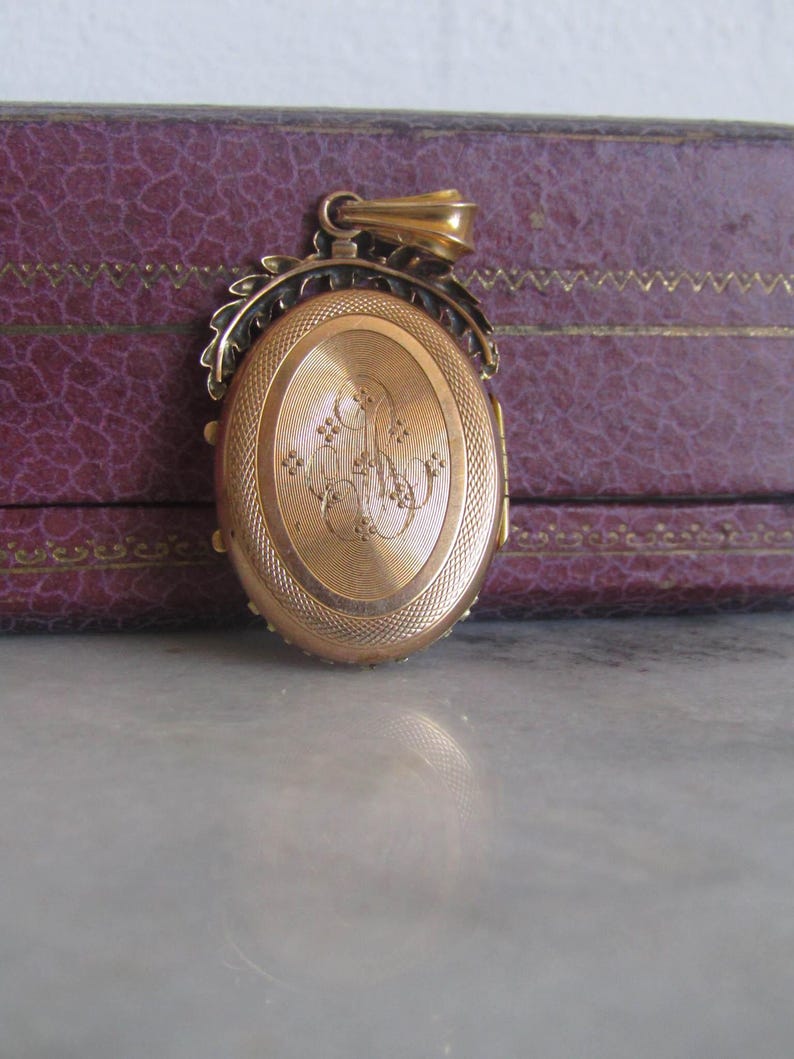 May include: A gold locket with an oval-shaped front and a detailed floral design around the edge. The front of the locket has an engraved monogram.
