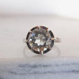 May include: A silver ring featuring a large, faceted, clear gemstone. The gemstone is round and set in a silver band with prongs. The ring is displayed on a white marble surface, showcasing its brilliance and design.