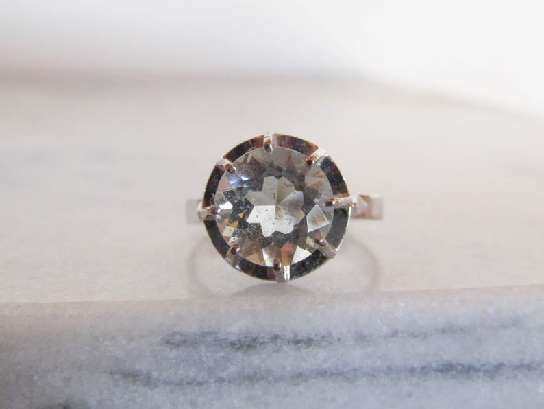 May include: A silver ring featuring a large, faceted, clear gemstone. The gemstone is round and set in a silver band with prongs. The ring is displayed on a white marble surface, showcasing its brilliance and design.