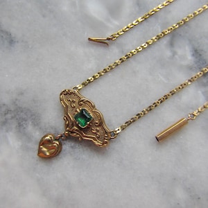 May include: A gold-toned necklace with an ornate pendant featuring a square green gemstone and a heart-shaped charm. The necklace has a delicate chain and is displayed on a marble surface.
