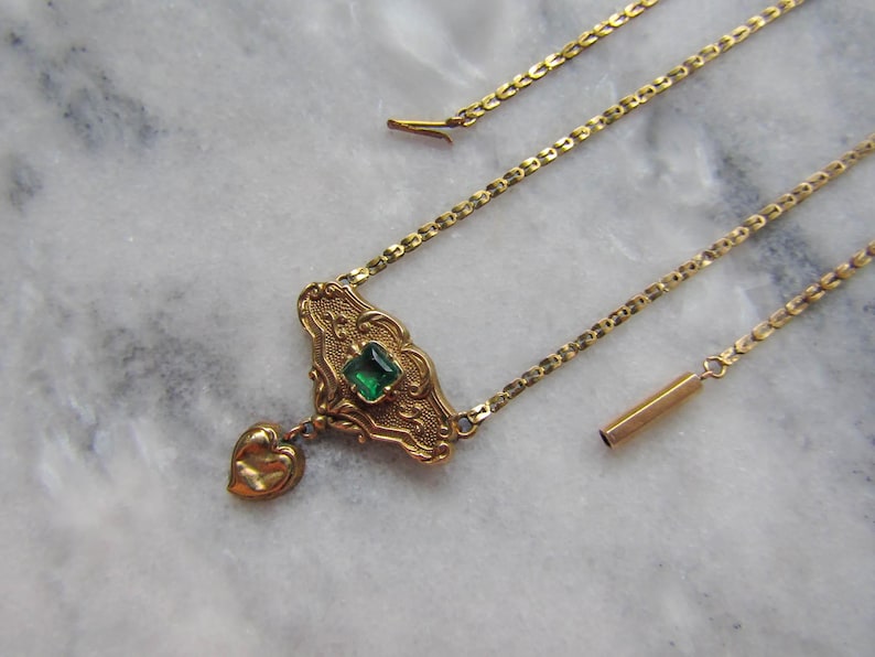 May include: A gold-toned necklace with an ornate pendant featuring a square green gemstone and a heart-shaped charm. The necklace has a delicate chain and is displayed on a marble surface.