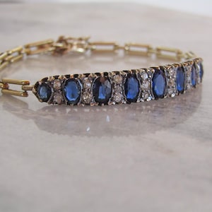 Op de afbeelding: Een goudkleurige armband met een rij ovale, diepblauwe edelstenen, afgewisseld met kleinere, heldere stenen. De armband heeft rechthoekige schakels. Waarschijnlijk een vintage of antiek sieraad.