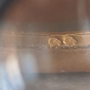 May include: Close-up of a gold ring with two small, engraved markings. The markings are difficult to read.