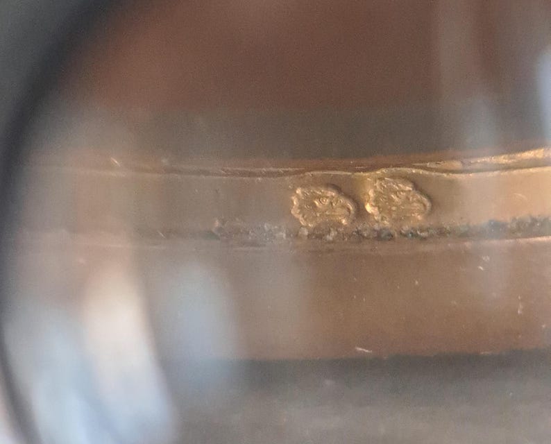 May include: Close-up of a gold ring with two small, engraved markings. The markings are difficult to read.
