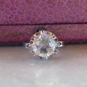 May include: A silver ring with a large, round, clear gemstone. The gemstone is set in a detailed, scalloped bezel. The ring is displayed on a light-colored surface, with a purple box in the background.