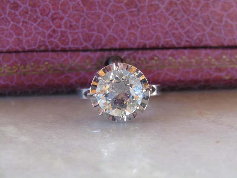 May include: A silver ring with a large, round, clear gemstone. The gemstone is set in a detailed, scalloped bezel. The ring is displayed on a light-colored surface, with a purple box in the background.