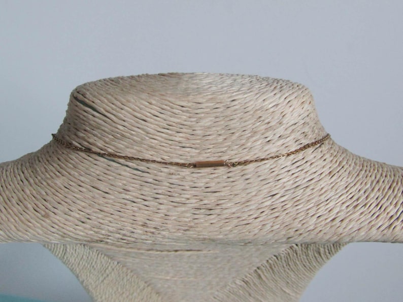 May include: A delicate gold-colored necklace displayed on a mannequin. The necklace features a fine chain with a small rectangular bar at the back. The mannequin is covered in a woven, beige material, set against a light blue background.