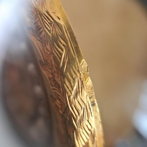 Op de afbeelding: Close-up van een goudkleurig, mogelijk antiek object met ingewikkelde gegraveerde patronen. Het ontwerp heeft een herhalend, getextureerd motief, mogelijk bloem- of bladachtig, waardoor een gedetailleerd oppervlak ontstaat. De rand van het object vertoont lichte slijtage.