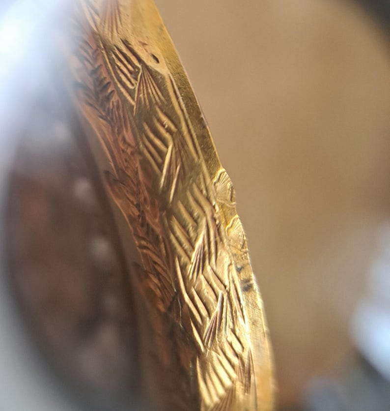 Op de afbeelding: Close-up van een goudkleurig, mogelijk antiek object met ingewikkelde gegraveerde patronen. Het ontwerp heeft een herhalend, getextureerd motief, mogelijk bloem- of bladachtig, waardoor een gedetailleerd oppervlak ontstaat. De rand van het object vertoont lichte slijtage.