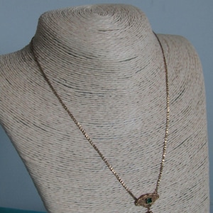 May include: A gold-colored necklace with a small pendant featuring a green gemstone and a heart-shaped charm. The necklace is displayed on a mannequin with a neutral-colored, textured surface, against a light blue background. The chain is delicate and the pendant is ornate.