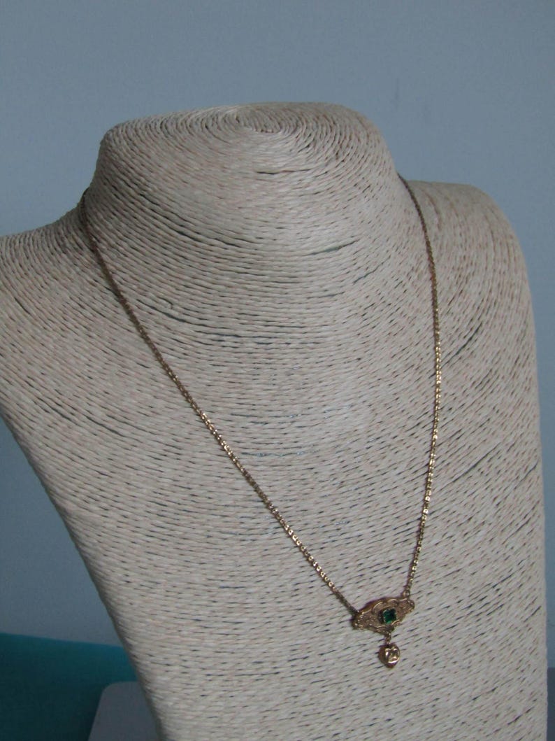 May include: A gold-colored necklace with a small pendant featuring a green gemstone and a heart-shaped charm. The necklace is displayed on a mannequin with a neutral-colored, textured surface, against a light blue background. The chain is delicate and the pendant is ornate.
