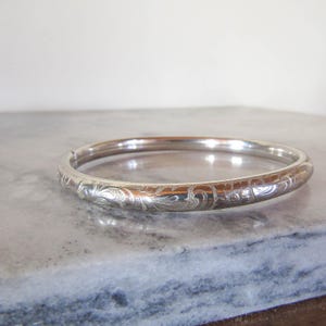 May include: A silver-toned metal bangle bracelet featuring a continuous engraved pattern of swirling floral and leaf motifs. The bracelet has a smooth, polished finish and a rounded profile, showcasing intricate detailing across its entire surface.