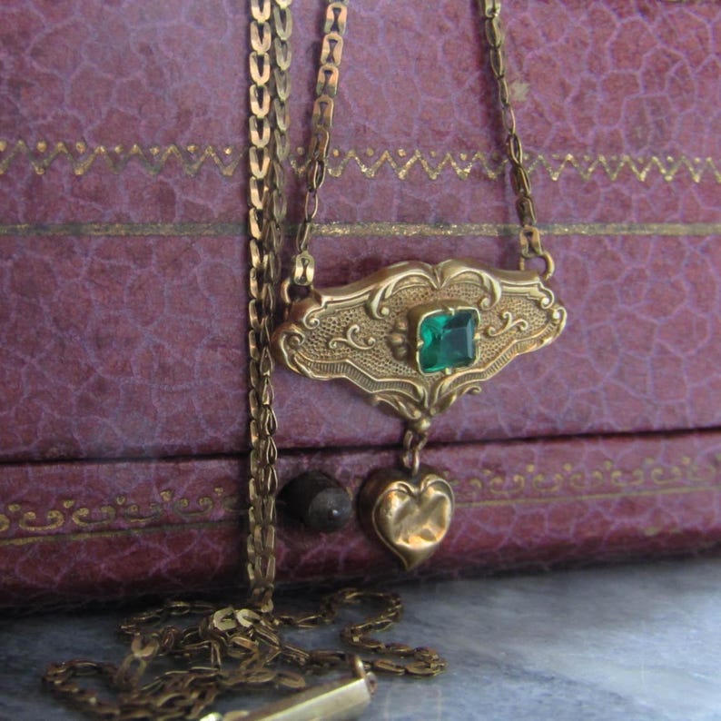 May include: A vintage gold-toned necklace with an ornate pendant featuring a square emerald-green gemstone. The pendant has a heart-shaped charm dangling below. The necklace is displayed against a textured, purple background.