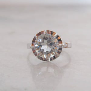 May include: A silver ring featuring a large, round, clear gemstone with a faceted cut. The gemstone is set in a decorative, multi-pronged setting. The ring is displayed on a white surface.