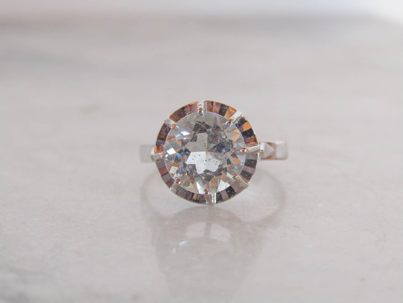 May include: A silver ring featuring a large, round, clear gemstone with a faceted cut. The gemstone is set in a decorative, multi-pronged setting. The ring is displayed on a white surface.