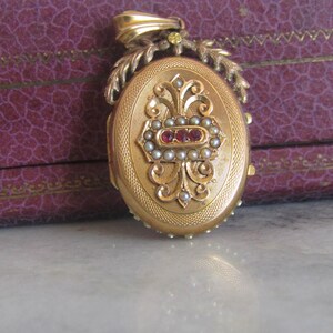 May include: A gold oval locket with a floral design and red and white gemstones. The locket is attached to a gold chain with a leaf design.