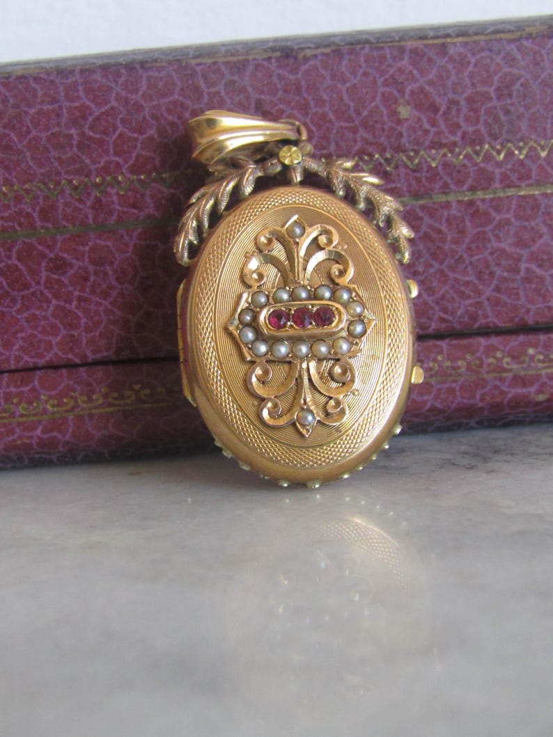 May include: A gold oval locket with a floral design and red and white gemstones. The locket is attached to a gold chain with a leaf design.