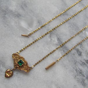 May include: A gold-toned necklace with an ornate pendant featuring a square green gemstone and a heart-shaped charm. The necklace has a delicate chain and a bar-shaped clasp. The jewelry is displayed on a marble surface.