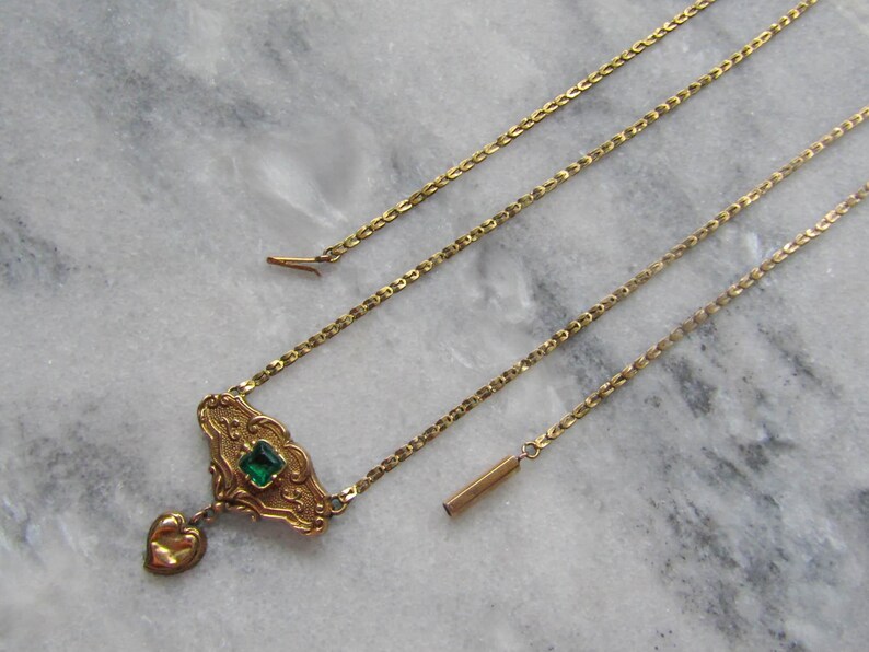 May include: A gold-toned necklace with an ornate pendant featuring a square green gemstone and a heart-shaped charm. The necklace has a delicate chain and a bar-shaped clasp. The jewelry is displayed on a marble surface.