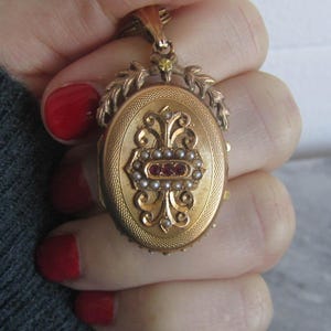 May include: A gold locket with a delicate floral design and a red and white stone accent. The locket is open and has a small mirror inside.