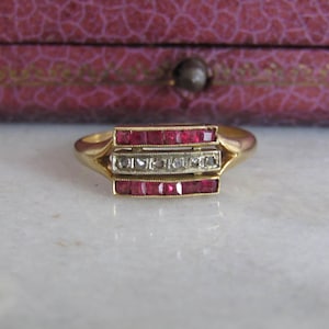 Puede incluir: Un anillo de oro con tres secciones rectangulares. Las secciones superior e inferior están engastadas con pequeñas piedras rojas. La sección central presenta una fila de piedras más pequeñas y transparentes. El anillo se muestra sobre un fondo borroso.