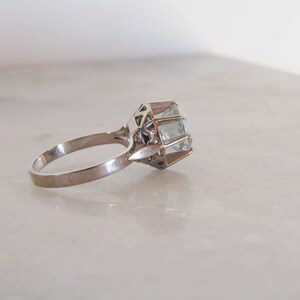 May include: A silver ring with a large, light blue gemstone. The gemstone is rectangular and set in a geometric, openwork setting. The ring is on a white surface.