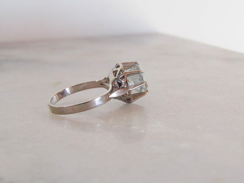 May include: A silver ring with a large, light blue gemstone. The gemstone is rectangular and set in a geometric, openwork setting. The ring is on a white surface.