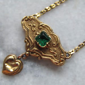 May include: A vintage gold-toned necklace featuring an ornate pendant with a square emerald-green gemstone and a small heart charm. The pendant is approximately 1.5 inches long and is suspended from a delicate chain.