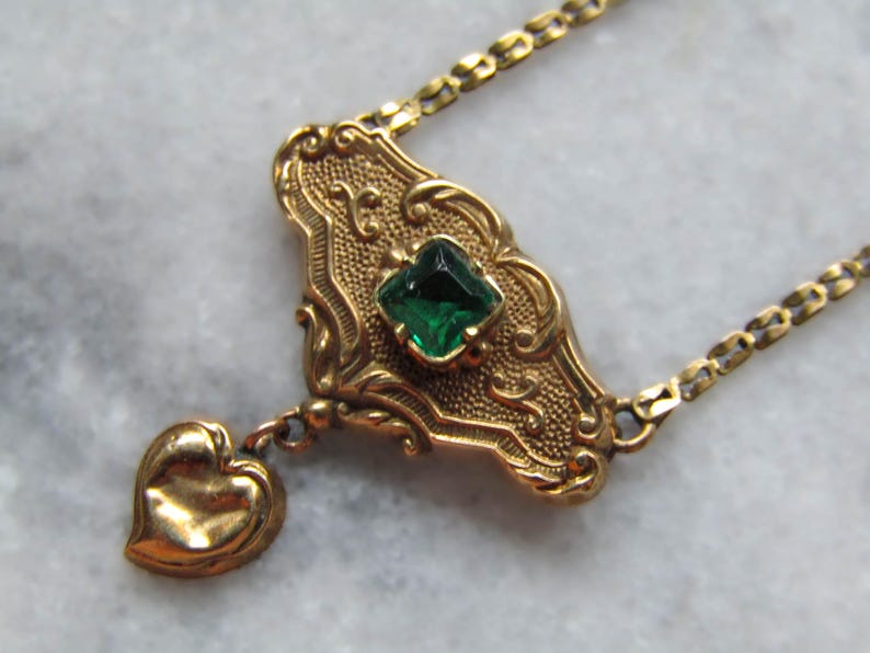 May include: A vintage gold-toned necklace featuring an ornate pendant with a square emerald-green gemstone and a small heart charm. The pendant is approximately 1.5 inches long and is suspended from a delicate chain.