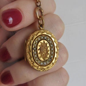 Op de afbeelding: Een goudkleurige, ovale locket met ingewikkelde bloemen- en bladerdetails. De locket heeft een centraal ontwerp omringd door kleine, ronde parels. Het hangt aan een fijne gouden ketting.