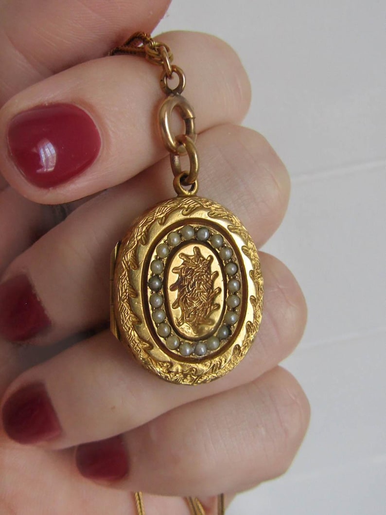 Op de afbeelding: Een goudkleurige, ovale locket met ingewikkelde bloemen- en bladerdetails. De locket heeft een centraal ontwerp omringd door kleine, ronde parels. Het hangt aan een fijne gouden ketting.