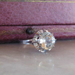 May include: A silver ring with a large, clear, faceted gemstone. The ring is set against a blurred background of a purple jewelry box. The gemstone is round and has many facets, reflecting light.