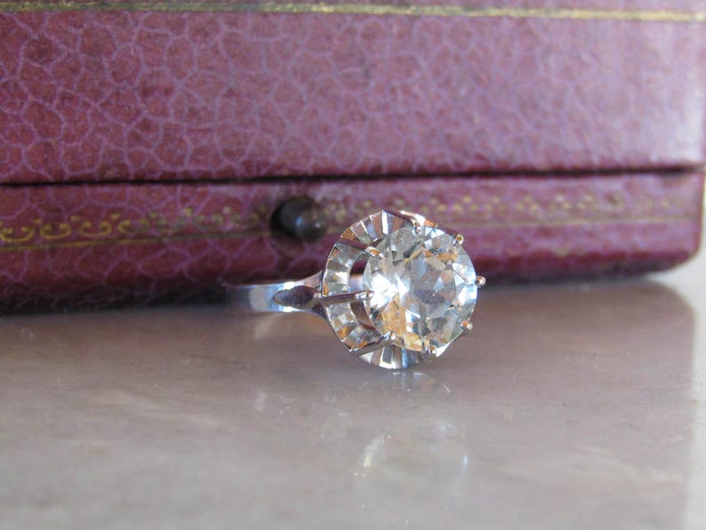 May include: A silver ring with a large, clear, faceted gemstone. The ring is set against a blurred background of a purple jewelry box. The gemstone is round and has many facets, reflecting light.