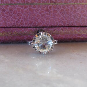 May include: A silver ring with a large, clear, round-cut gemstone. The gemstone is set in a detailed, scalloped bezel. The ring is displayed on a marble surface, with a purple jewelry box in the background.