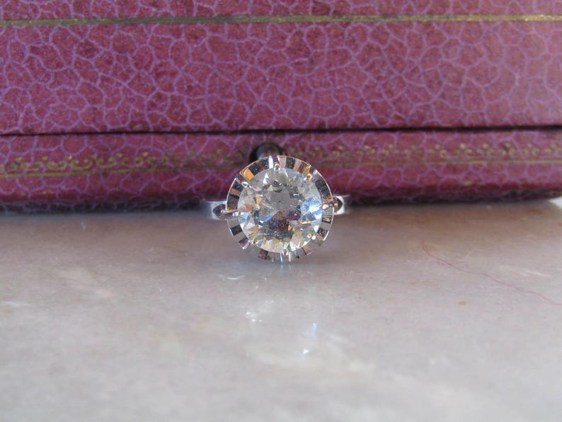May include: A silver ring with a large, clear, round-cut gemstone. The gemstone is set in a detailed, scalloped bezel. The ring is displayed on a marble surface, with a purple jewelry box in the background.