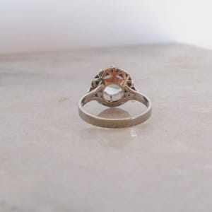 May include: A silver ring with a large, round, light-colored gemstone at the center. The gemstone is surrounded by a decorative, openwork setting. The ring is set against a neutral background.