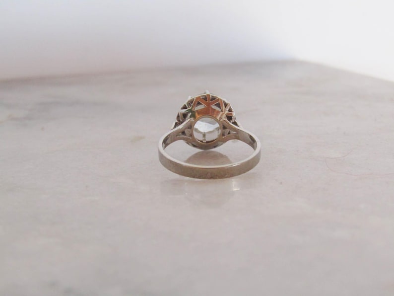 May include: A silver ring with a large, round, light-colored gemstone at the center. The gemstone is surrounded by a decorative, openwork setting. The ring is set against a neutral background.