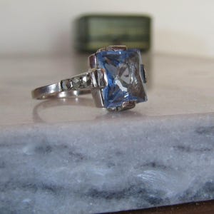 Könnte beinhalten: Ein silberfarbener Ring mit einem großen, rechteckigen, facettierten hellblauen Stein in einer Krappenfassung. Die Ringschiene ist auf jeder Seite mit drei kleinen, runden, dunkelgrauen Steinen verziert, die zur zentralen Fassung führen.