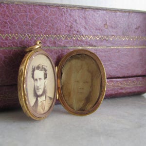Op de afbeelding: Open gouden medaillon met twee ovale compartimenten, elk met een sepia-getint portret. E&eacute;n toont een man in pak en baard. Het medaillon ligt in de buurt van een paarse, getextureerde doos.